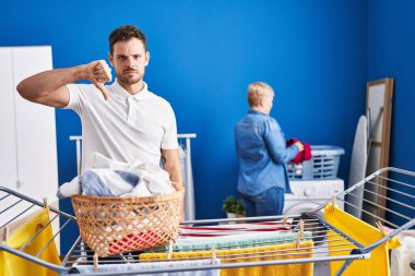 Hispanic mother and son hanging clothes at clothesline with angry face, negative sign showing dislike with thumbs down, rejection concept 