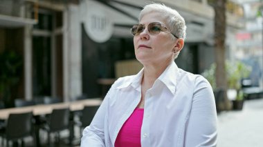 Orta yaşlı, gri saçlı, güneş gözlüklü, kahve dükkanının terasına gülümseyen bir kadın.