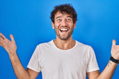 Hispanic young man standing over blue background celebrating crazy and amazed for success with arms raised and open eyes screaming excited. winner concept 