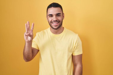 Young hispanic man standing over yellow background showing and pointing up with fingers number three while smiling confident and happy. 