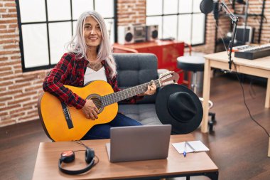 Müzik stüdyosunda dizüstü bilgisayar kullanarak klasik gitar çalan gri saçlı kadın müzisyen.
