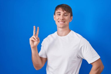 Mavi arka planda dikilmiş, gülümseyen, parmaklarını zafer işareti yaparken gösteren beyaz, sarışın bir adam. İki numara.. 