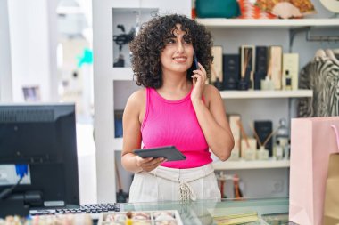 Orta Doğulu genç bir bayan mağaza asistanı dokunmatik ped kullanıyor ve elbise mağazasında akıllı telefondan konuşuyor.