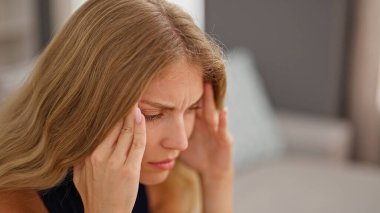 Evdeki kanepede oturan genç sarışın kadın baş ağrısı çekiyor.