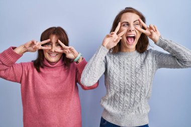 Anne ve kızı mavi arka planda duruyorlar barış sembolü yapıyorlar parmakları yüzünde, neşeyle gülüyorlar zaferi gösteriyorlar. 