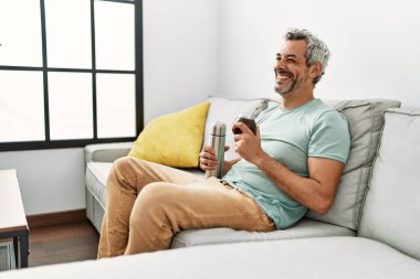 Middle age grey-haired man drinking mate infusion sitting on sofa at home