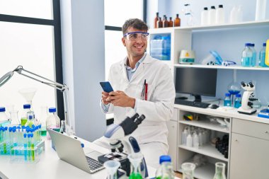 Young hispanic man scientist smiling confident using smartphone at laboratory