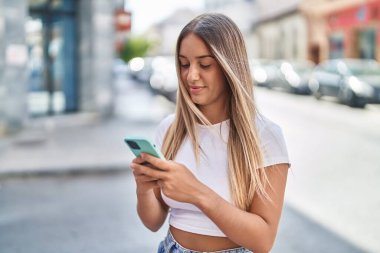 Genç, güzel, İspanyol bir kadın sokakta akıllı telefon kullanarak gülümsüyor.