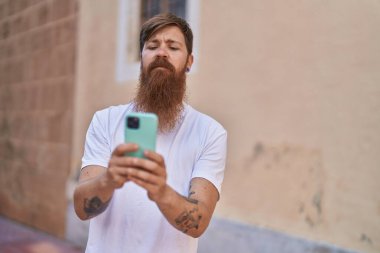 Genç kızıl saçlı adam sokakta rahat bir ifadeyle akıllı telefon kullanıyor.