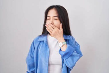 Beyaz arka planda duran genç Çinli kadın ağzıyla ağzını kapatmaktan sıkılmış. Huzursuz ve uykulu.. 