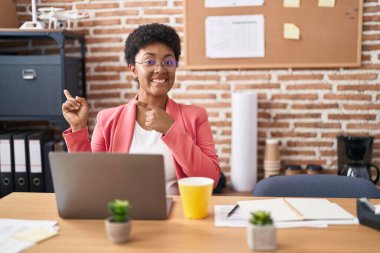 Ofiste çalışan Afro-Amerikalı genç bir kadın. Gözlüklü, gülümseyen, kameraya bakan ve iki eliyle ve parmaklarıyla onu işaret eden.. 