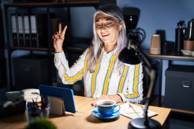 Gri saçlı, gece ofiste çalışan orta yaşlı bir kadın kameraya bakıp gülümseyen ve zafer işareti yapan. İki numara.. 