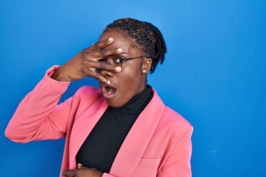 Güzel siyah kadın mavi arka planda dikiliyor. Şoka giriyor. Yüzü ve gözleri kapanıyor, utangaç ifadesiyle parmaklarının arasından bakıyor.. 