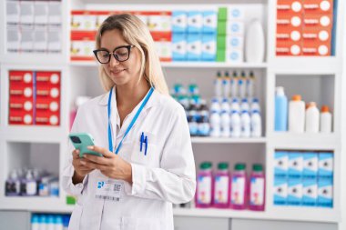 Young blonde woman pharmacist using smartphone working at pharmacy