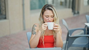 Kahve dükkanının terasında laptopla kahve içen genç sarışın kadın.