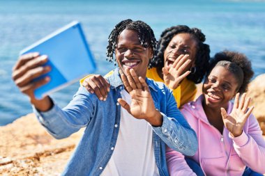 African american friends having video call sitting on rock at seaside