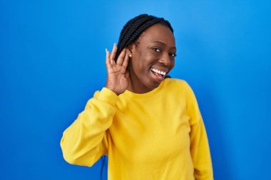 Beautiful black woman standing over blue background smiling with hand over ear listening an hearing to rumor or gossip. deafness concept. 