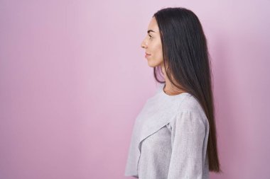 Genç esmer kadın pembe arka planda duruyor. Yan tarafa bakıyor, rahat poz veriyor. Kendine güvenen bir gülümsemesi var.. 