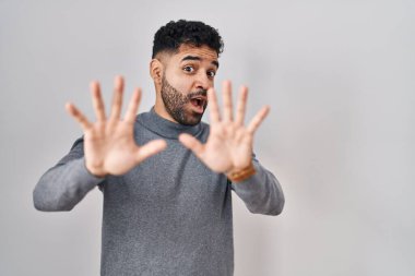 Hispanic man with beard standing over white background afraid and terrified with fear expression stop gesture with hands, shouting in shock. panic concept. 