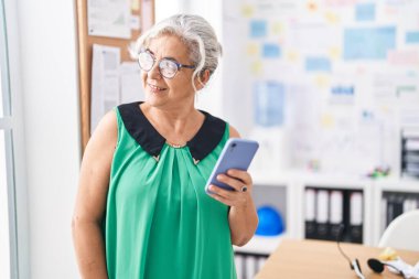 Orta yaşlı, gri saçlı, iş kadını. Ofiste akıllı telefon kullanarak gülümsüyor.