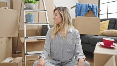 Genç sarışın kadın yeni evinde yerde kendinden emin bir şekilde gülümsüyor.