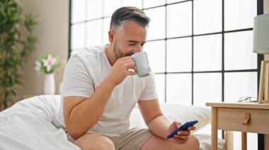 Beyaz saçlı adam akıllı telefon kullanıyor. Yatak odasında kahve içiyor.