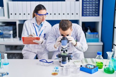 Laboratuardaki panoya mikroskop kullanan kadın ve erkek bilim adamları yazıyor.