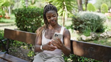 Parkta bankta oturan Afro-Amerikan kadın akıllı telefon kullanıyor.
