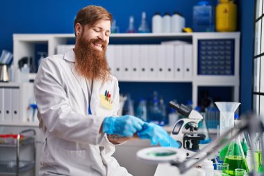 Genç kızıl saçlı bilim adamı gülümsüyor. Laboratuvarda eldiven takıyor.