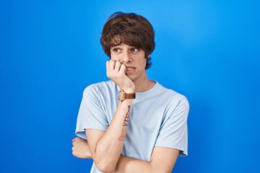Hispanic young man standing over blue background looking stressed and nervous with hands on mouth biting nails. anxiety problem. 
