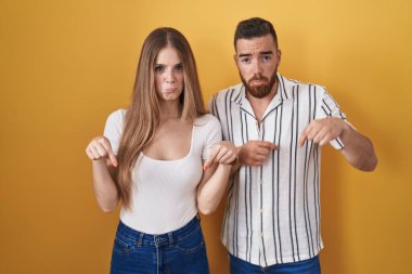 Sarı arka planda duran genç çift üzgün ve üzgün görünüyor, parmaklarıyla yön gösteriyor, mutsuz ve depresif.. 