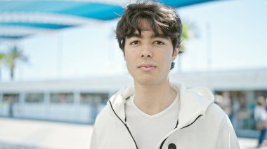 Young hispanic man standing with serious expression at park