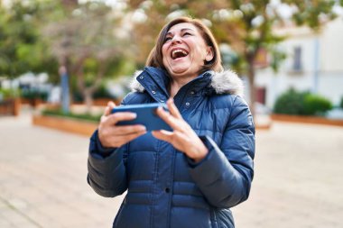 Orta yaşlı kadın parkta akıllı telefondan video izlerken kendinden emin bir şekilde gülümsüyor.