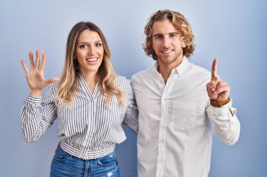 Mavi arka planda duran genç çift, altı numaralı parmaklarıyla gülümseyerek mutlu ve kendinden emin bir şekilde işaret ediyor.. 