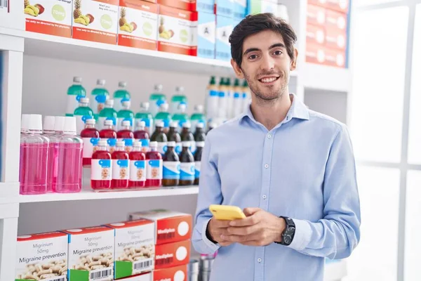 Young hispanic man customer smiling confident using smartphone at pharmacy