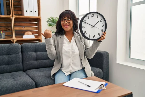 Hispanic woman working at therapy office holding clock pointing thumb up to the side smiling happy with open mouth