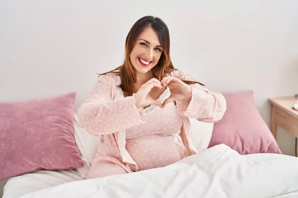 Young pregnant woman doing heart gesture with hands sitting on bed at bedroom