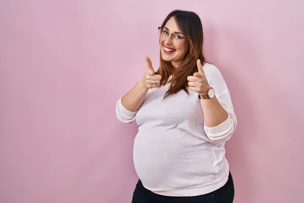 怀孕的女人站在粉色的背景上 手指指向镜头 脸上洋溢着欢乐和滑稽的表情 充沛的精力和活力 — 图库照片