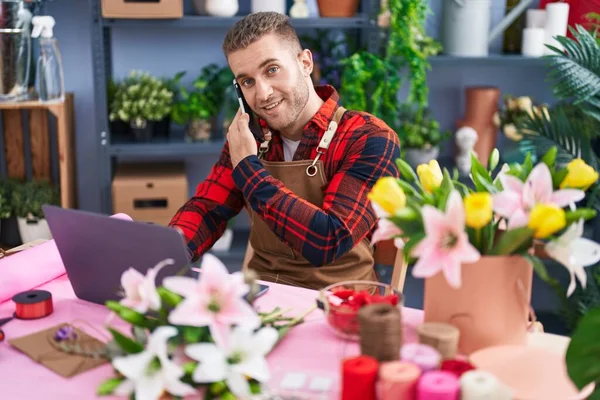 Young caucasian man florist talking on smartphone using laptop at flower shop