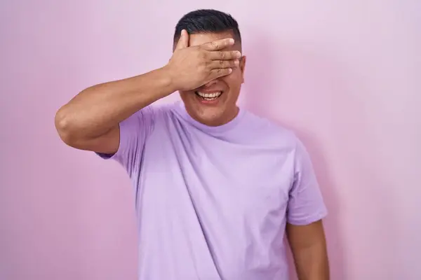 ピンクの背景の上に立つ若いヒスパニック系の男が笑顔で 驚きのために目をカバー顔に手で笑っています 視覚障害の概念 — ストック写真