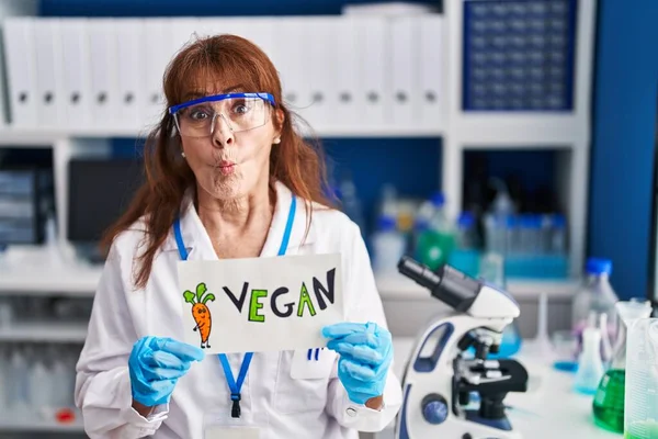 Middle age hispanic woman working at vegan laboratory making fish face with mouth and squinting eyes, crazy and comical.