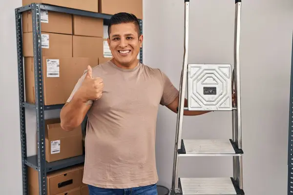 Hispanic young man working at small business ecommerce holding ladders smiling happy and positive, thumb up doing excellent and approval sign