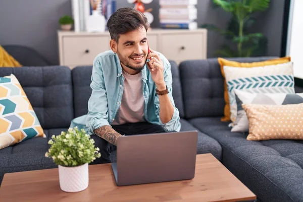 Young hispanic man talking on smartphone using laptop at home