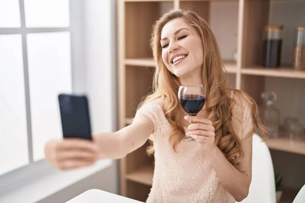 Young blonde woman drinking wine making selfie by the smartphone at home