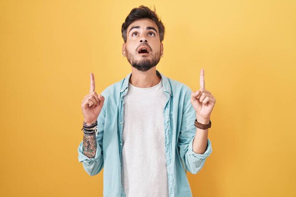 Young hispanic man with tattoos standing over yellow background amazed and surprised looking up and pointing with fingers and raised arms. 