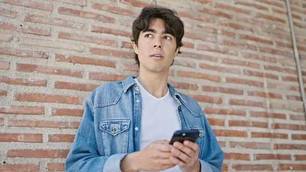 Genç İspanyol adam, izole tuğla arka planda ciddi bir ifadeyle akıllı telefon kullanıyor.