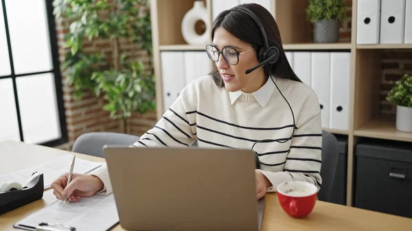 Young beautiful hispanic woman call center agent having video call writing on document at office