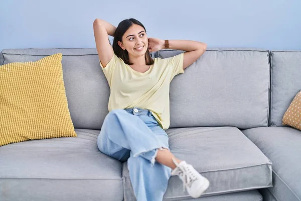 Young hispanic woman relaxed with hands on head sitting on sofa at home