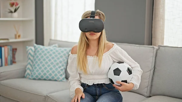 Young blonde woman watching soccer match using virtual reality glasses at home