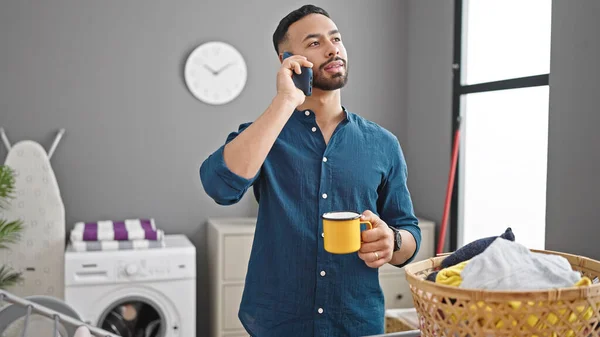 Young hispanic man talking on smartphone drinking coffee waiting for washing machine at laundry room